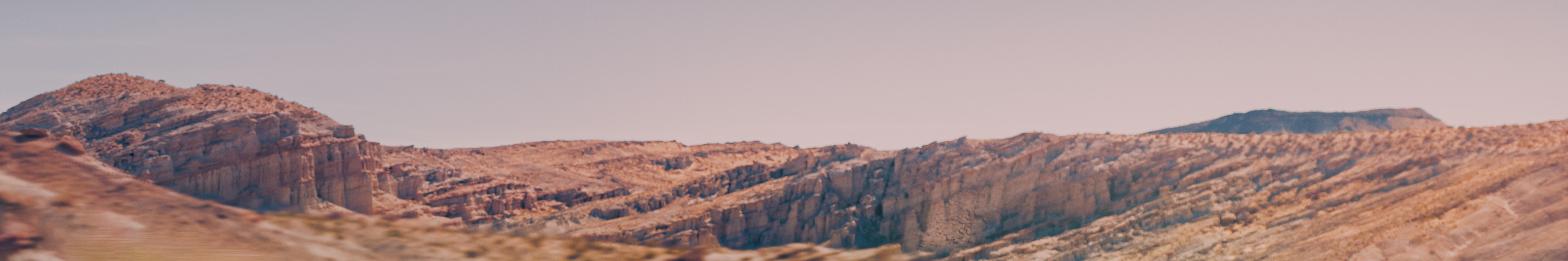 Wide, sweeping panoramic view of layers of red and tan rock formations in a vast, arid desert landscape under a pale sky