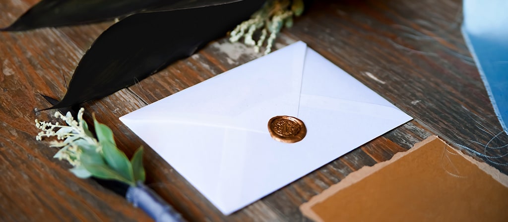 An envelope with a decorative seal holding an invitation lays on a wooden surface with floral arrangements