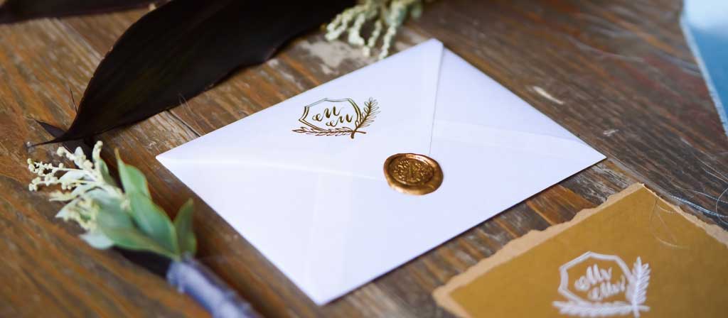 An envelope with a decorative seal holding an invitation lays on a wooden surface with floral arrangements