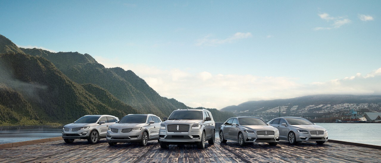 A number of Lincoln vehicles in the Platinum White exterior color are shown parked near a lake and a range of mountains