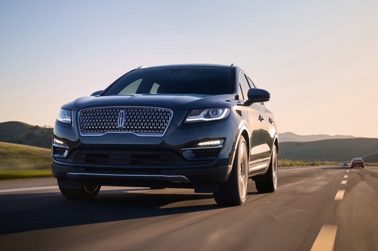 A Lincoln M K C is being shown in the Black Velvet exterior color driving through a country road