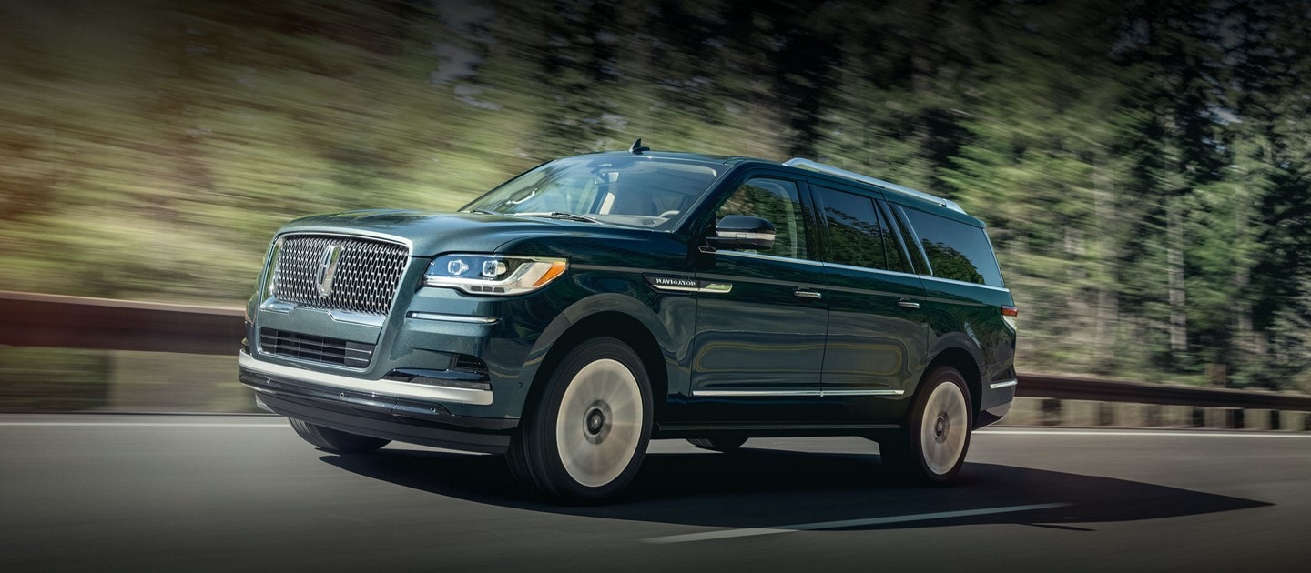 A 2024 Lincoln Navigator® SUV in Flight Blue is being driven around a curve surrounded by greenery