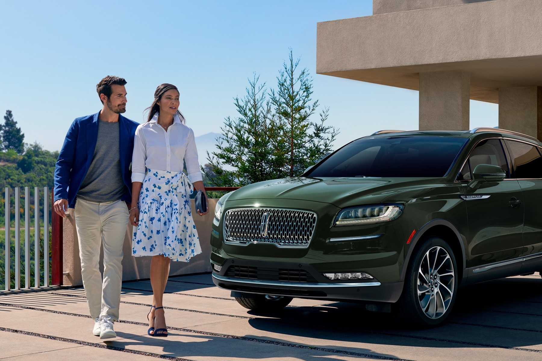 A couple walk around the front of a green Lincoln Aviator SUV parked in the driveway of a modern home near the mountains.