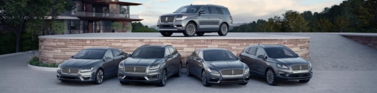 Various Lincoln 2019 vehicles in gray or silver parked in showcase near trees and large house