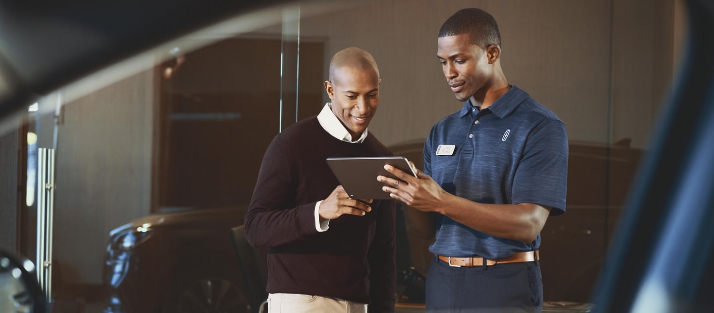 A man wearing a nametag and a Lincoln logo polo shirt shows information on a tablet computer to another man, as seen through the window of a Lincoln SUV