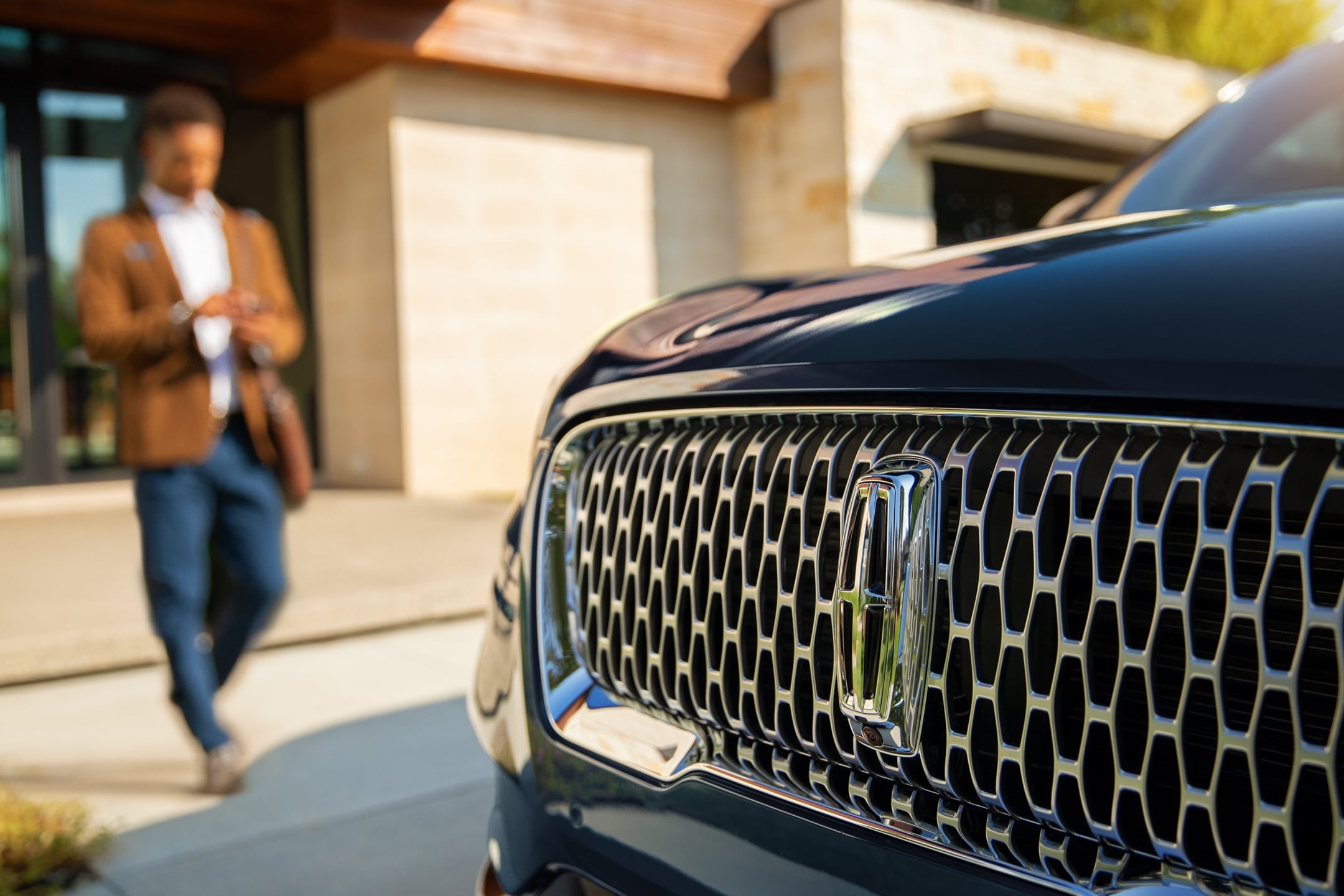A close-up of the grill of a Lincoln Corsair SUV. In the background, a man approaches the car from the front door of a home
