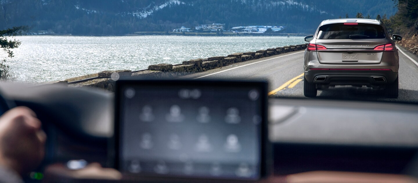 View through 2023 Lincoln Aviator® windshield of another vehicle being driven ahead on a road next to a mountain lake