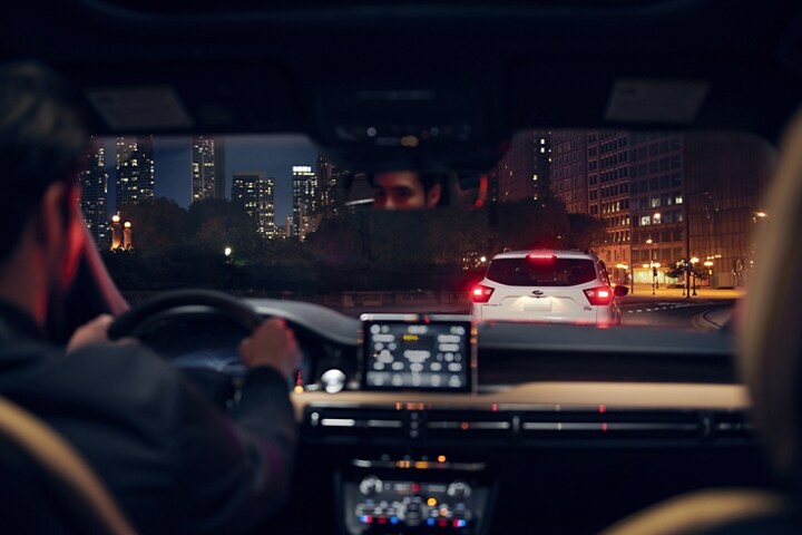 Through the windshield of a Lincoln Corsair we see a vehicle with its brake lights illuminated ahead on a city street with a skyline in the distance
