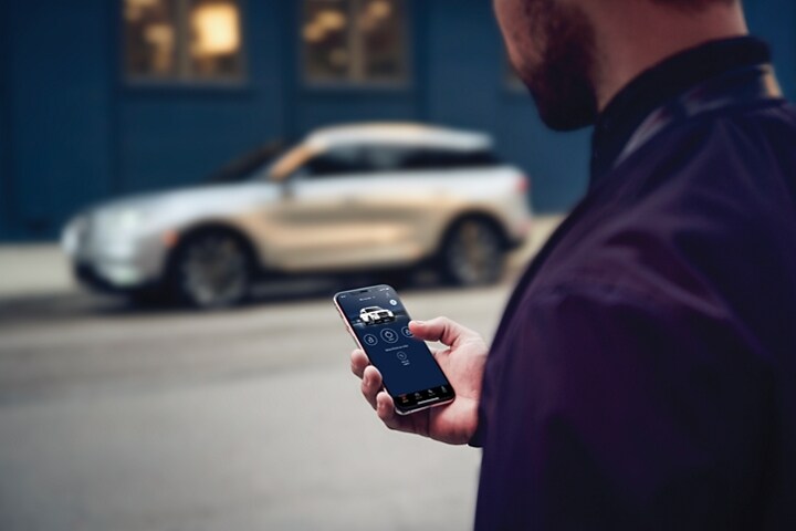 A man is standing across the street from his Lincoln vehicle interacting with the Lincoln way app on his smart phone device