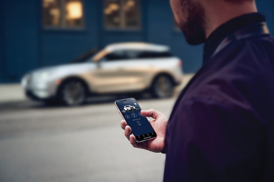 A man is standing across the street from his Lincoln vehicle interacting with the Lincoln Way App on his smartphone device.