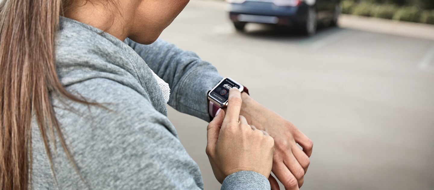 A woman is standing by a car in a parking lot during the day interacting with a smartwatch on her wrist using Lincoln Connect