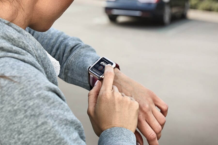 A woman is standing by a car in a parking lot during the day interacting with a smart watch on her wrist using Lincoln Connect