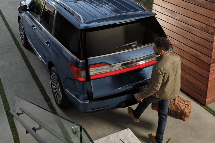 A man carrying a brown duffel bag and phone is kicking his foot under the rear bumper of a 2021 Lincoln Navigator to open the hands free lift gate