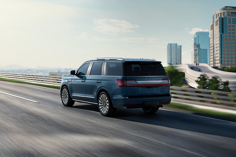 A 2021 Lincoln Navigator in Blue Diamond is being driven on a freeway against the blur of sunlit greenery and a cityscape