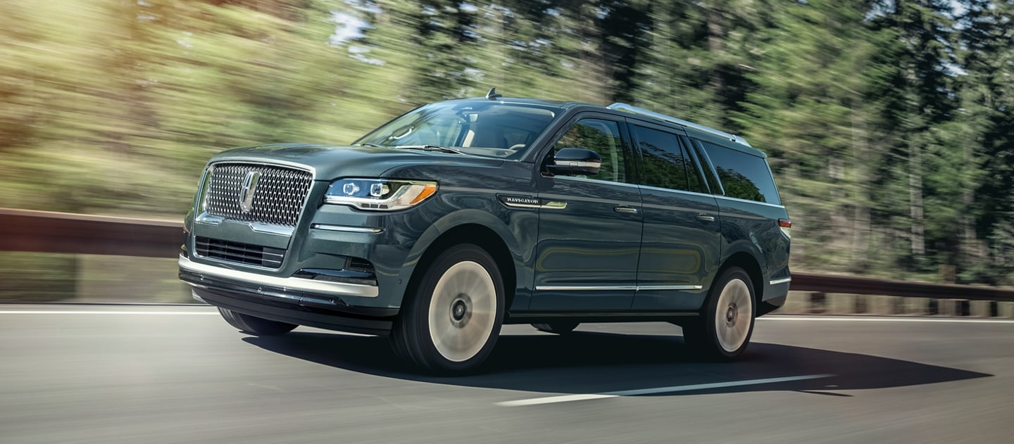 A 2023 Lincoln Navigator® SUV is being driven on a highway lined with tall pine trees and bathed in sunlight