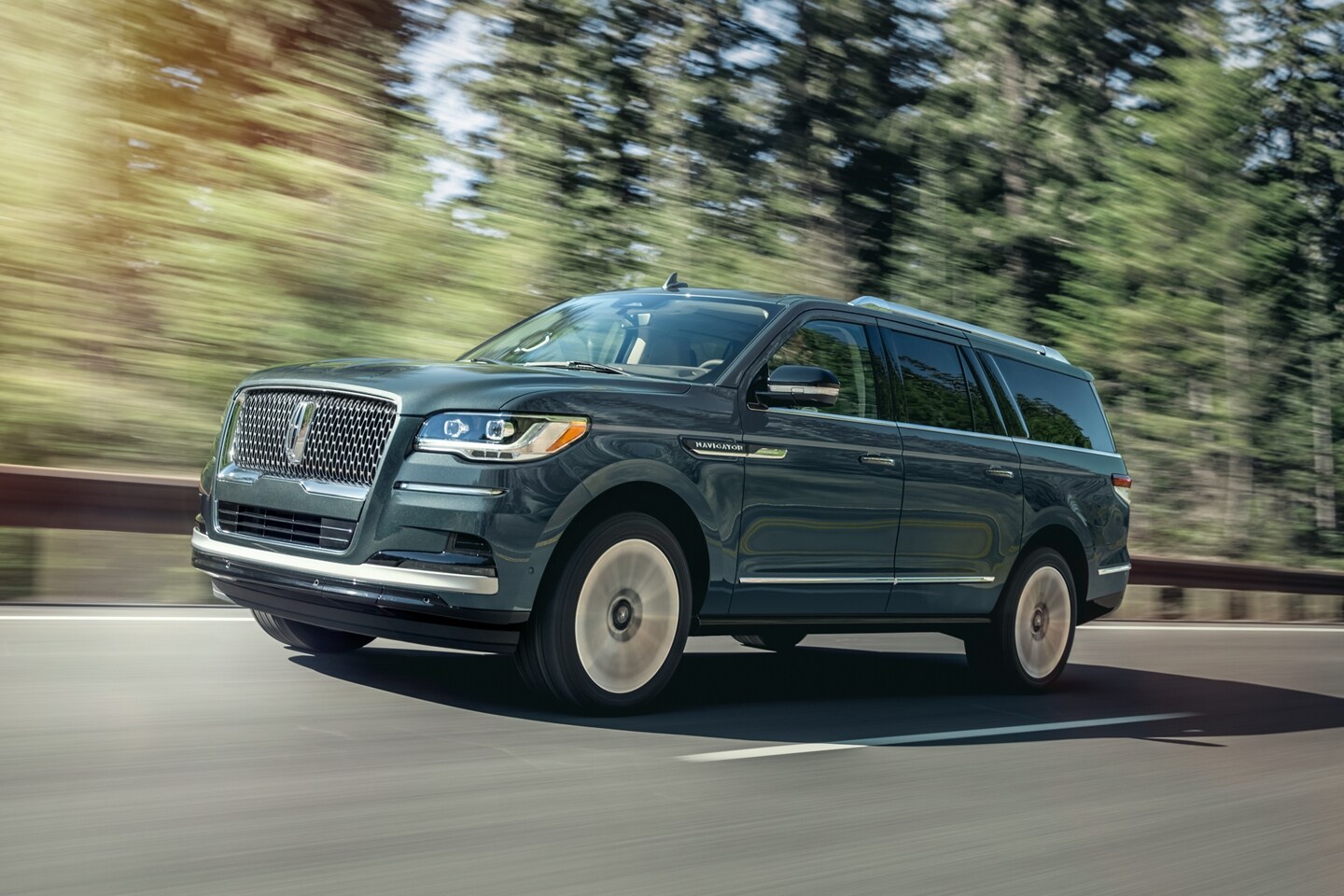 A 2023 Lincoln Navigator SUV is being driven on a highway lined with tall pine trees as it bathes in sunlight