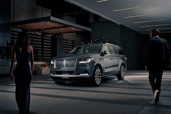 A man and woman in formal wear are approaching a 2023 Lincoln Navigator SUV at night as it lights with the Lincoln Embrace