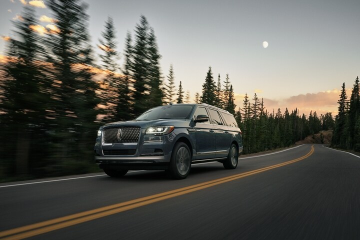 A 2023 Lincoln Navigator SUV is being driven along a road lined with tall pine trees with a faint moon in the sky at dusk
