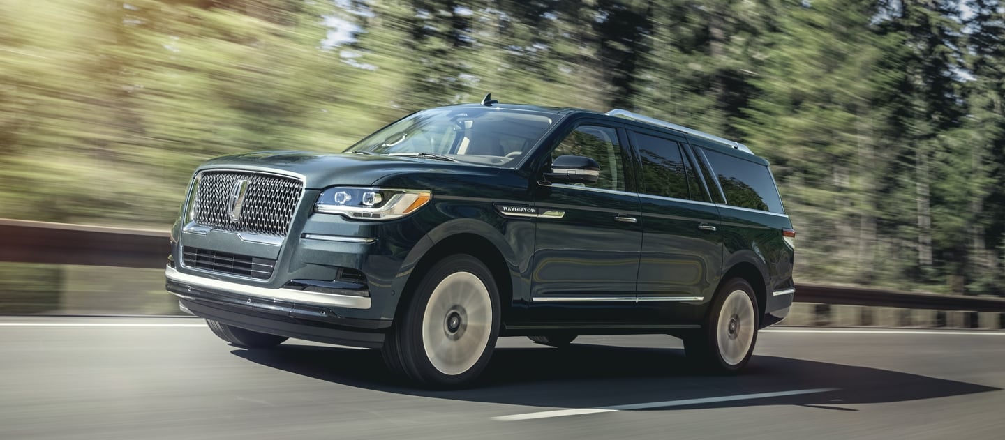 A 2024 Lincoln Navigator SUV is being driven on a highway lined with tall pine trees and bathed in sunlight