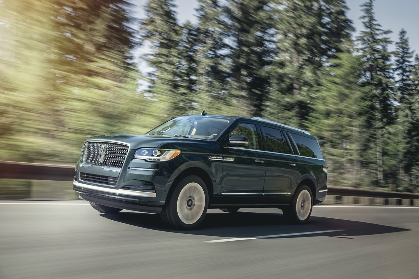 A 2024 Lincoln Navigator SUV is being driven on a highway lined with tall pine trees as it bathes in sunlight