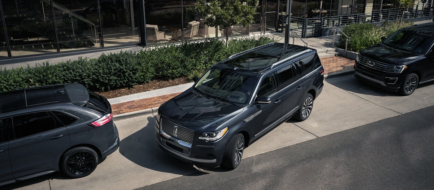 A 2024 Lincoln Navigator SUV is being parallel parked between two cars on a downtown road outside a luxury hotel
