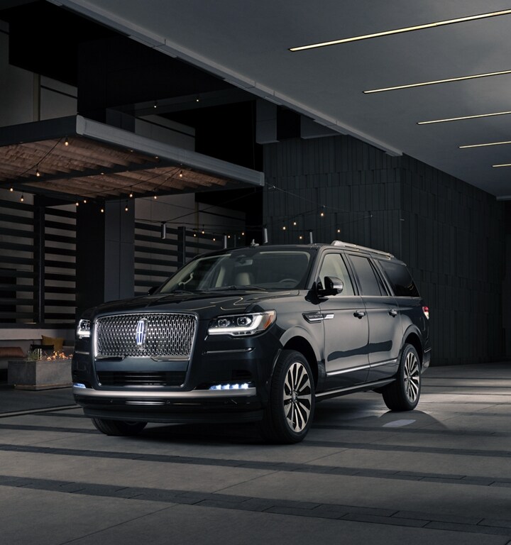 A man and woman in formal wear are approaching a 2024 Lincoln Navigator SUV at night as it lights with the Lincoln Embrace