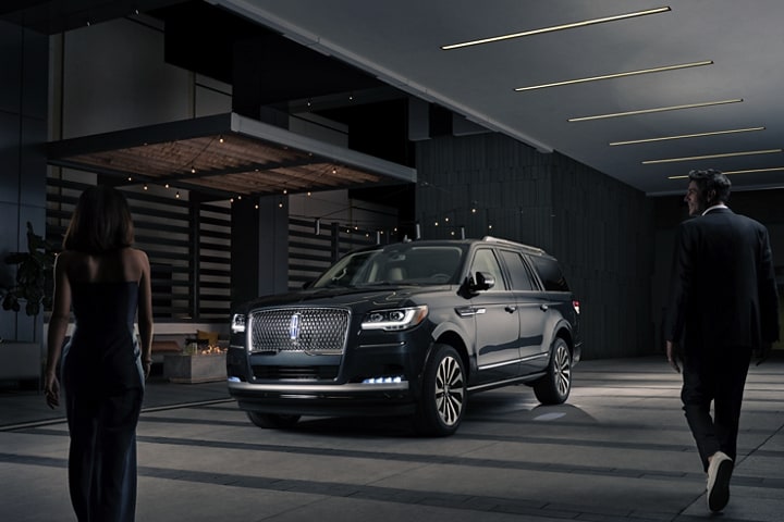 A man and woman in formal wear are approaching a 2024 Lincoln Navigator SUV at night as it lights with the Lincoln Embrace