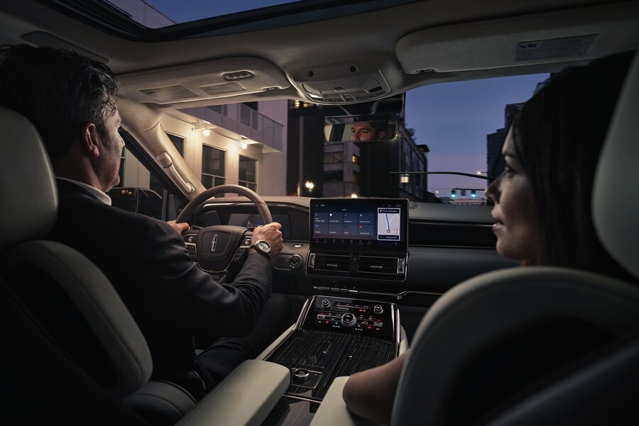 A man and woman are relaxing inside a 2024 Lincoln Navigator SUV as they drive through a downtown street at night