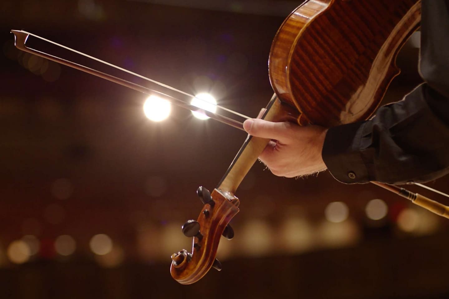 The bow of a musician’s violin slides across stage spotlights to illustrate the dramatic sound of the symphonic chimes